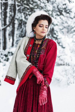 Foto de Atractiva mujer morena con maquillaje de noche, en un traje de estilo boho étnico rojo con bordado, chaqueta blanca, sombrero de piel, guantes de cuero rojo, falda en un parque forestal nevado de invierno - Imagen libre de derechos