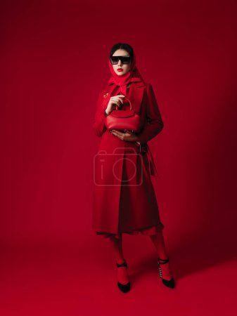 Téléchargez les photos : Total look rouge style mode studio portrait d'une magnifique jeune femme brune, manteau, sac, lunettes, foulard, maquillage, broche à plumes, lèvres rouges, talons hauts - en image libre de droit