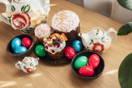 Téléchargez les photos : Assiettes avec trois gâteaux de Pâques orthodoxes traditionnels et des œufs rouges, verts et bleus peints, et théières de fleurs vintage se tiennent sur la table de fête en bois - en image libre de droit