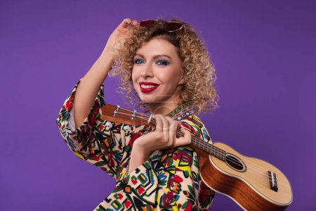 Photo for Beautiful blonde woman with red lips and curly hair, in the red glasses, stands on a purple studio background, holds an ukulele in her hands and smiles - Royalty Free Image