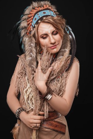 Téléchargez les photos : Belle femme blonde avec maquillage, vêtue dans un style ethnique indien, avec un cafard sur la tête, avec un bouquet d'épillets dans les mains, se tient sur un fond de studio noir et sourit - en image libre de droit