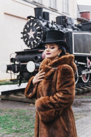 Foto de Retro retrato de estilo de hermosa mujer morena en traje vintage y sombrero de copa cerca del tren viejo, abrigo de piel, trenzas de color, maquillaje de noche, ojos azules - Imagen libre de derechos