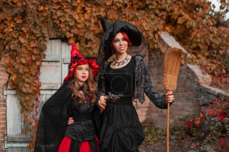 Téléchargez les photos : Deux sorcières, mère et fille, dans les robes noires et rouges et chapeaux pointus, avec un balai, debout devant la vieille maison aux feuilles jaunes, costumes d'Halloween - en image libre de droit