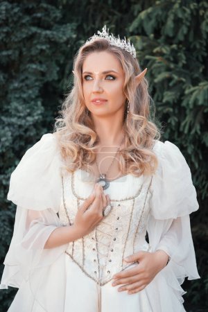 Foto de Hermosa mujer rubia elfa con ojos azules, en un vestido de estilo medieval blanco y en una corona, está de pie en flores blancas, princesa de fantasía - Imagen libre de derechos