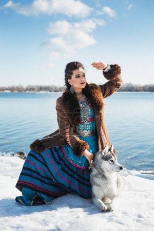 Photo for Beautiful brunette woman with grey husky dog is sitting on the snow, braid hairstyle, blue and pink boho ethnic style costume, brown fur coat, evening makeup, silver accessories, winter, lake, sky - Royalty Free Image