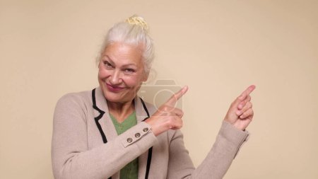 Photo for An caucasian elderly woman points her finger to attract attention on a beige background - Royalty Free Image