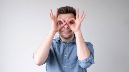 Photo for A young European man holds his fingers to his eyes like binoculars, as if looking into the distance. Studio shot - Royalty Free Image
