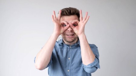 Téléchargez les photos : Un jeune Européen tient ses doigts sur ses yeux comme des jumelles, comme s'il regardait au loin. Plan studio - en image libre de droit