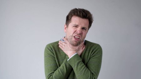El joven europeo está sufriendo de dolor de garganta, irritación, y agarra su garganta debido a sensaciones desagradables. Captura de estudio
