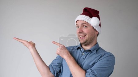 Der fröhliche junge Mann mit dem Neujahrsmütze deutet auf einen leeren Raum und präsentiert ein Produkt. Studioaufnahme