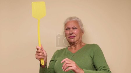An elderly European woman is trying to catch a mosquito with a fly swatter. Studio shot