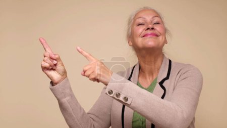 Une femme âgée caucasienne pointe du doigt pour attirer l'attention sur un fond beige