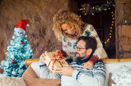 Photo for Happy couple enjoy christmas sharing gifts leisure activity at home. Xmas decorations and tree in background. Man receiving a surprise box present from his wife woman behind. People and gift xmas - Royalty Free Image