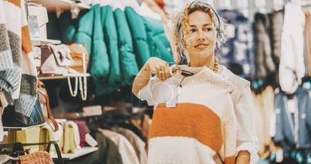 Photo for Woman doing shopping activity inside a garment clothes store, smiling and looking sweaters. Sales holiday discount season on commercial center. Female people and fashion trendy business. Happy lady - Royalty Free Image