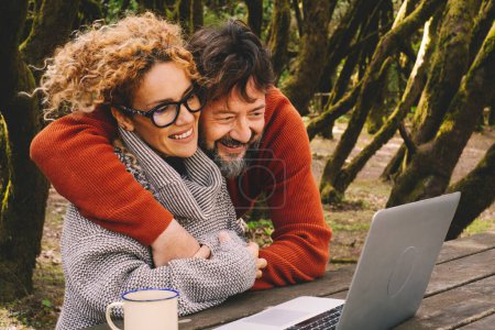  glückliches Paar im Freien mit Laptop auf einem Holztisch mit Wald im Hintergrund. Mann und Frau genießen die Technologie und die roaming Internet-Verbindung nach draußen. Paar bei Videoanruf am Computer