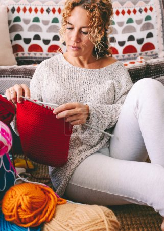 Téléchargez les photos : Portrait de scène de la vie réelle de la jeune femme d'âge moyen qui fait du tricot à la maison assise sur le sol. Les gens et les loisirs. Loisirs d'intérieur femme tricot laine rouge seul. Une personne à la main fait un travail - en image libre de droit
