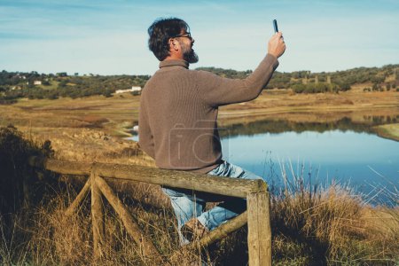 Téléchargez les photos : Touriste masculin prendre une photo de paysage en utilisant un téléphone mobile avec vue sur le lac. Un homme profite d'une activité de loisirs de plein air au parc et reste connecté à une connexion Internet en itinérance sur un appareil cellulaire - en image libre de droit