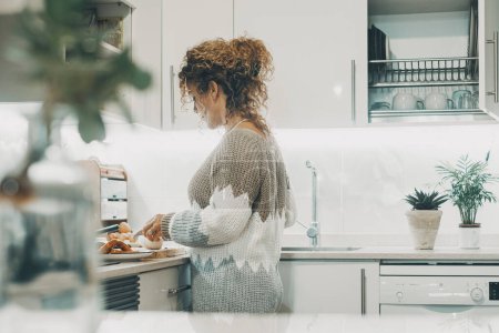 Téléchargez les photos : Vue latérale réelle de la cuisine femme à la maison dans la cuisine moderne minimale blanche. Une femme cuisine seule dans les loisirs d'intérieur. Femme ou dame indépendante jouissant de son temps à l'intérieur - en image libre de droit