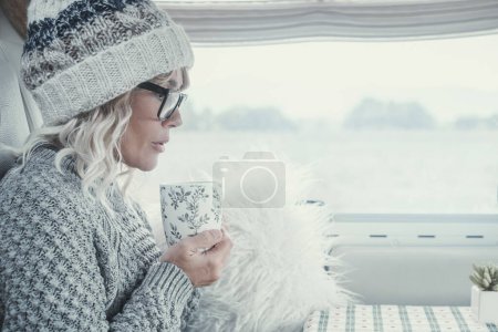 Foto de Vista lateral de una atractiva dama con el pelo blanco y ropa bebiendo de una taza dentro de su caravana durante la temporada de invierno. Imagen brillante de la mujer serena. Retrato de mujeres en el ocio interior - Imagen libre de derechos