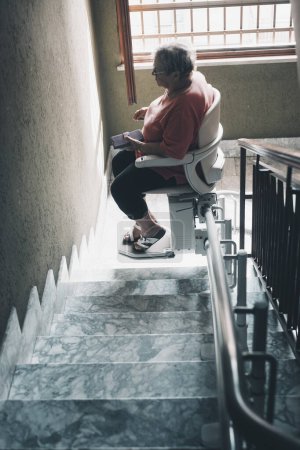 Téléchargez les photos : Vieille femme avec des problèmes de jambes utilisent chaise moteur électrique pour monter les escaliers à la maison dans un bâtiment. Technologie pour aider les personnes âgées concept - en image libre de droit