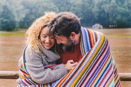 Téléchargez les photos : Couple d'adultes amoureux romantique étreinte avec tendresse sous une couverture de laine colorée dans un parc extérieur. Concept d'activité de loisirs d'hiver pour les hommes et les femmes en relation. Personnes embrassant et appréciant - en image libre de droit