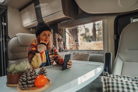 Foto de Mujer que vive dentro de una caravana moderna casa rodante sentado a la mesa y el uso de un teléfono móvil para mantenerse conectado. Tecnología de conexión itinerante para viajeros mujeres en estilo de vida van - Imagen libre de derechos