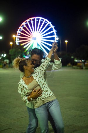 Téléchargez les photos : Couple de jeunes adultes dansent en plein air avec des attractions Luna Park en arrière-plan. Concept de célébration et de bonheur avec l'homme et la femme s'amuser ensemble dans la nuit en plein air. Des gens joyeux célèbrent - en image libre de droit