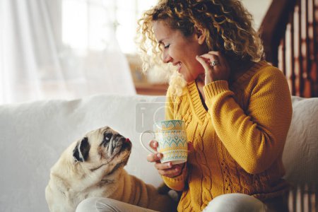 Smiling woman looking with love her dog best friend having relax leisure indoor activity sitting on the sofa. Concept of people and animals lifestyle. Mature female and pug enjoy home together