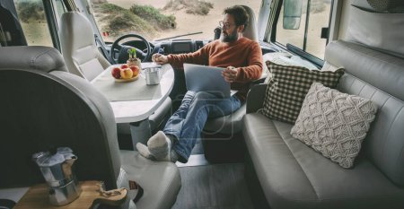 One alone man sitting and relaxing inside a camper van alternative home vanlife lifestyle off grid using a laptop and taking a coffee in total relax freedom. Indoor motor home travel leisure activity