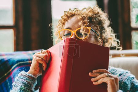 Photo for Woman relax and pleasure expression about reading a book novel alone at home. Happiness and joyful emotion in female people face portrait. Book cover on her face. Indoor leisure activity alone at home - Royalty Free Image