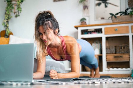 Photo for Healthy sport fitness lifestyle at home using laptop to watch and follow online lessons. Cheerful woman on the floor doing push ups plank exercise to build a perfect healthy body alone. Workout people - Royalty Free Image
