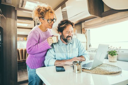 Happy couple man and woman working and using together a laptop computer inside a camper van planning holiday vacation destination and living vanlife lifestyle in freedom. People travel and tourism