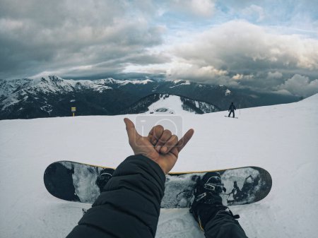 Téléchargez les photos : Point of view of man in waves hello sign with hand relaxing sitting on the snow in ski facilities and using snowboard to ride and enjoy active sport lifestyle and winter mountains vacation alone - en image libre de droit