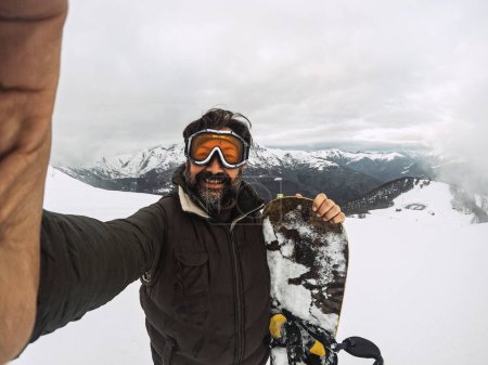 Téléchargez les photos : Mature adult man taking selfie picture in snow ski facilities with snowboard like young boy. People smiling at the camera and enjoying winter holiday mountain vacation. Senior rider - en image libre de droit