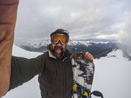 Téléchargez les photos : Selfie portrait of old mature man with snowboard enjoying winter holiday vacation alone. Youthful senior people in young sport activity. Enjoying outdoor leisure activity at the mountains with snow - en image libre de droit