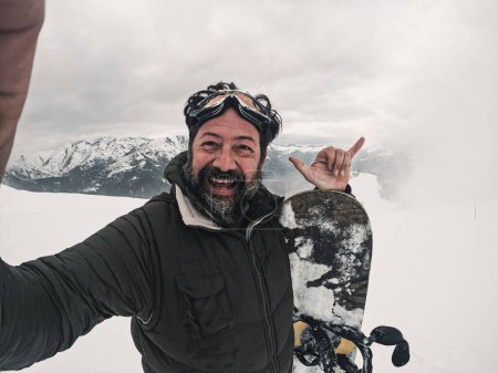 Photo for Mature adult man having fun and enjoying snowboard in winter holiday vacation taking selfie picture with action cam or phone. Tourist on snow in ski facilities. Mature adult rider enjoying outdoor - Royalty Free Image