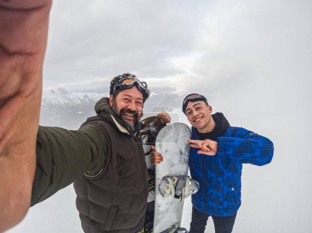 Téléchargez les photos : Father and son in friendship enjoying together winter holiday vacation doing snowboard and taking selfie picture with phone. Young and mature smiling and having fun in outdoor mountains leisure - en image libre de droit