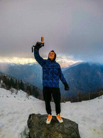 Téléchargez les photos : Young boy taking selfie footage with action cam of him standing and smiling with great mountain view in background. Concept of modern lifestyle and social media travel content sharing - en image libre de droit