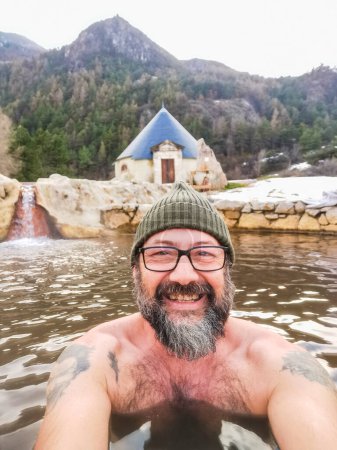 Foto de Selfie picture of happy man having fun in warm thermal outdoors water alone. Snow and mountains in background. People enjoying natural spa water with winter and mountains in background - Imagen libre de derechos