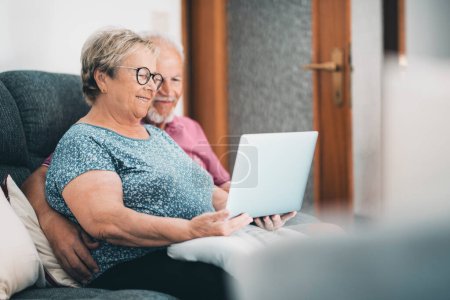 Senior couple at home having relax using laptop together. New modern lifestyle for mature retired people. Man and woman with computer and internet connection sitting on the sofa in indoor leisure