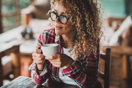 Foto de Attractive middle age woman with long curly hair enjoying relax at home drinking tea or coffee alone sitting at the table. Serene female people portrait smiling and thinking. Concept of single life - Imagen libre de derechos