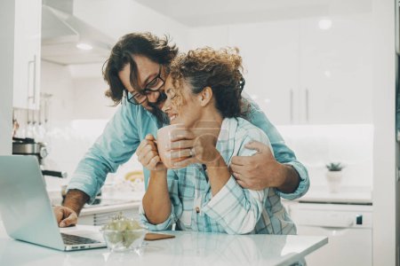 Foto de Man hugging woman at home in the kitchen with love. Indoor leisure activity and relationship lifestyle. Married people enjoying time. Young couple using computer and have romantic moment in real life - Imagen libre de derechos