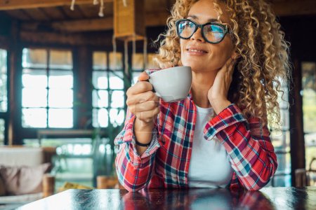 Foto de Serene woman at home drinking tea or coffee and enjoying relax leisure indoor activity alone sitting at the table. Portrait of cheerful and happy female people smiling and wearing eyewear. Curly hair - Imagen libre de derechos