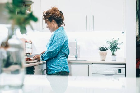 Foto de Indoor cooking leisure activity with happy young mature woman preparing food for lunch or dinner at home in white minimal kitchen alone. Living alone real life lifestyle female people middle age - Imagen libre de derechos