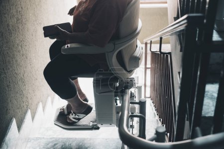 Foto de Aged woman needs modern assistance to go upstairs at home. Age problems concept. An electric, motorized chair life for persons with disabilities on a carpeted staircase in a residential home. - Imagen libre de derechos