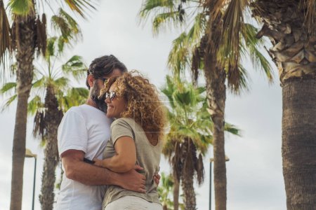 Téléchargez les photos : Adult couple man and woman enjoying relationship hugging with happiness outdoor in a palms street during summer holiday travel vacation. Outdoor leisure activity people in love and friendship smiling - en image libre de droit