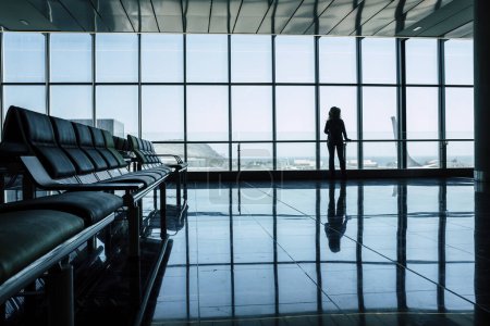 Téléchargez les photos : Travel concept. Back view of woman passenger in silhouette waiting her flight at the gate. Blue modern light mood. Tourist wait to fly. Business or vacation trip. Enjoying airplane to transport - en image libre de droit
