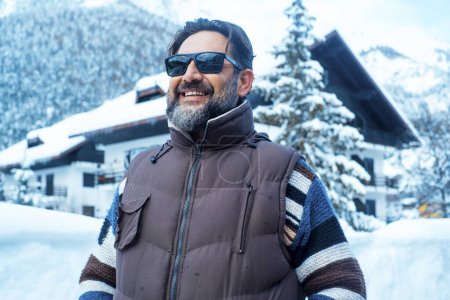 Foto de Portrait of a standing man smiling in winter snowfall, scenic place. Wooden home chalet and mountains in background. Snowfall. Happy cheerful male people in winter holiday vacation travel lifestyle - Imagen libre de derechos