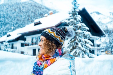 Foto de Side portrait of happy adult woman smiling and enjoying snow mountain outdoor leisure activity alone. Beautiful wood chalet house in background with trees pine in the garden outside. Winter holiday - Imagen libre de derechos