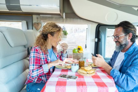 Foto de Mature man and woman enjoying time inside a modern camper van eating together and talking. Happy couple in vehicle travel activity. Using phone during brunch. Freedom adventure life people - Imagen libre de derechos